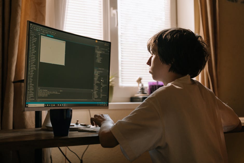 A person with short, dark hair wearing a white shirt is seated at a wooden desk in a room with natural light coming through beige blinds. They are working on a computer with a large monitor displaying
